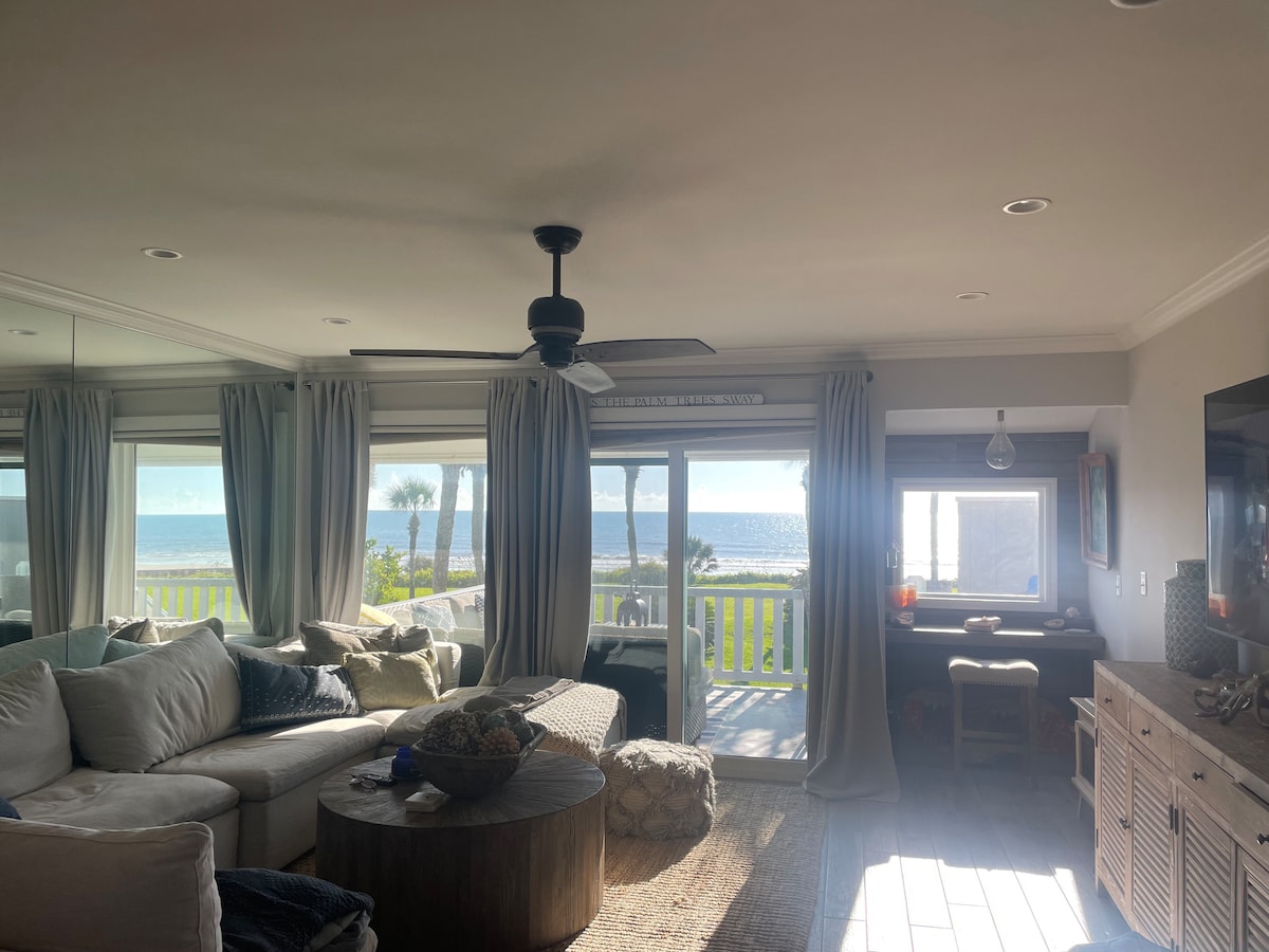 A bright living area features large windows showcasing ocean views. A spacious sectional sofa is arranged around a central coffee table. Soft, light curtains frame the windows, allowing natural light to fill the space. A work desk is visible in the background.
