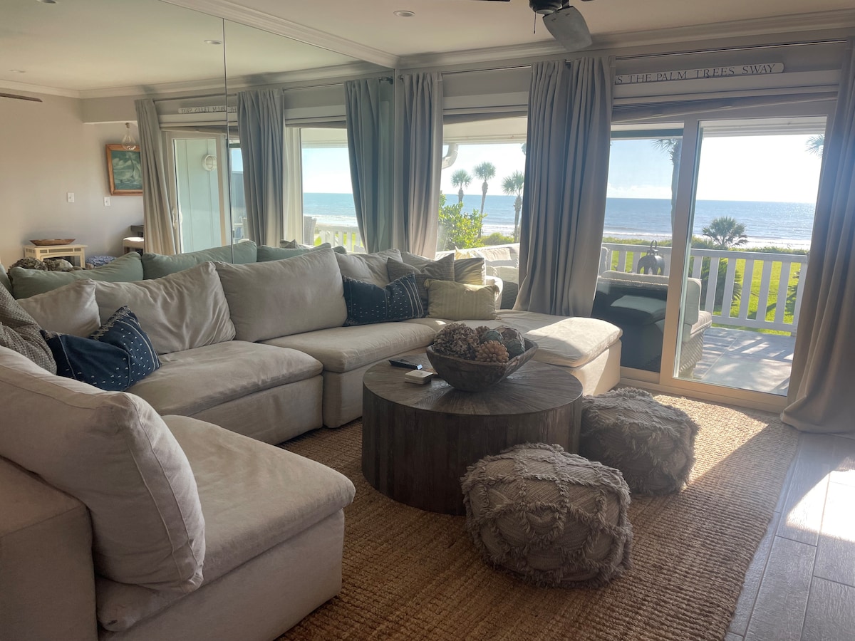A spacious living area is furnished with a large sectional sofa, complemented by textured poufs. Natural light floods the space through sliding glass doors, revealing views of the ocean and palm trees beyond. A round wooden coffee table adds a rustic touch to the cozy setting.