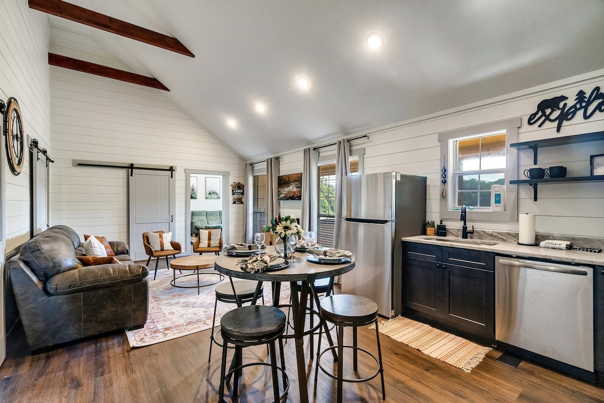 An open-concept living area features a modern kitchen with stainless steel appliances and a dining table set for four. A cozy seating area includes a brown sofa and two armchairs arranged around a circular coffee table. Large windows allow natural light to fill the space.
