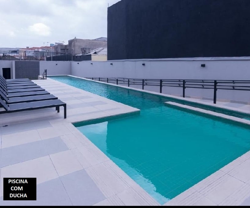 A rectangular swimming pool is visible, featuring clear turquoise water. Alongside the pool, several lounge chairs are arranged. A shower area is indicated nearby, and the surrounding space is paved with light-colored tiles. The background shows a neutral-colored wall and a cloudy sky.