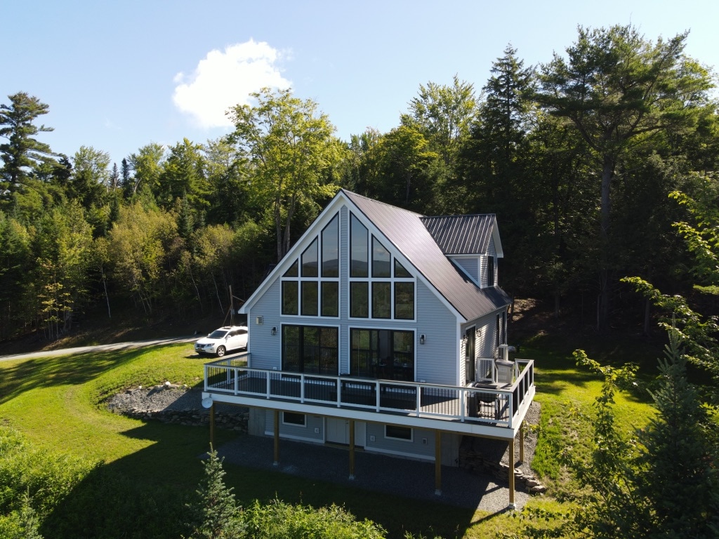 A modern chalet is showcased, featuring a unique A-frame design with large windows and a spacious deck. Surrounded by lush greenery, the property sits amidst a serene landscape, with a gravel driveway leading to the house and a parked vehicle visible.