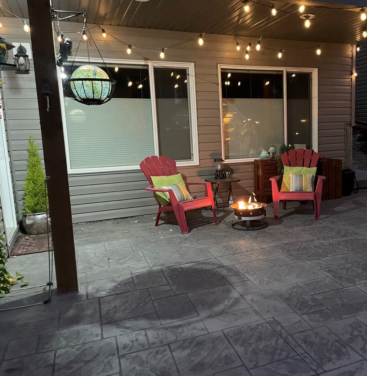 An inviting outdoor patio features two bright red Adirondack chairs positioned around a small fire pit. Warm string lights hang above, creating a cozy ambiance. The grey stone flooring complements the relaxed atmosphere, while large windows provide a view into the home.