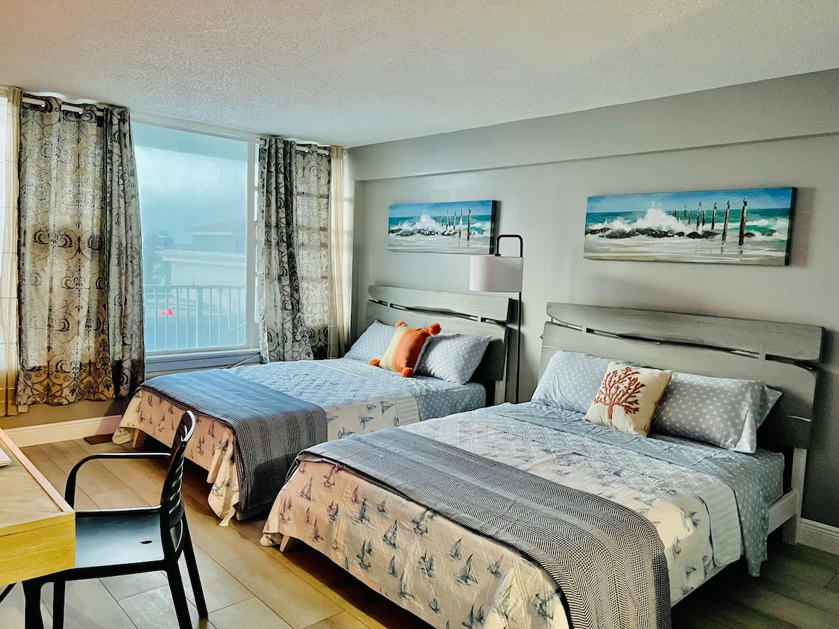 A bright and airy guest room features two spacious beds covered in light linens, flanked by decorative pillows. Large windows allow natural light to fill the space, while elegant curtains frame the views. A small table and chair are positioned near the window.