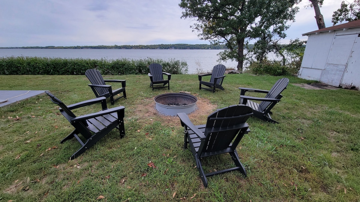 A circle of six black Adirondack chairs surrounds a fire pit on a grassy area by the lake. The tranquil water is visible in the background, bordered by lush greenery. The scene promotes an inviting space for relaxation and gatherings.