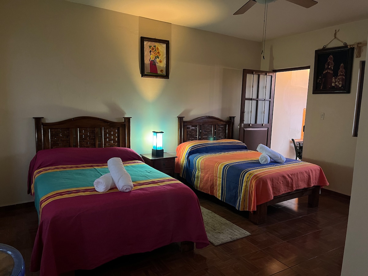 The room features two spacious beds, each adorned with colorful blankets. Light from a bedside lamp adds a soft glow, while art pieces decorate the walls. A doorway leads to another area, enhancing the room's functional layout.
