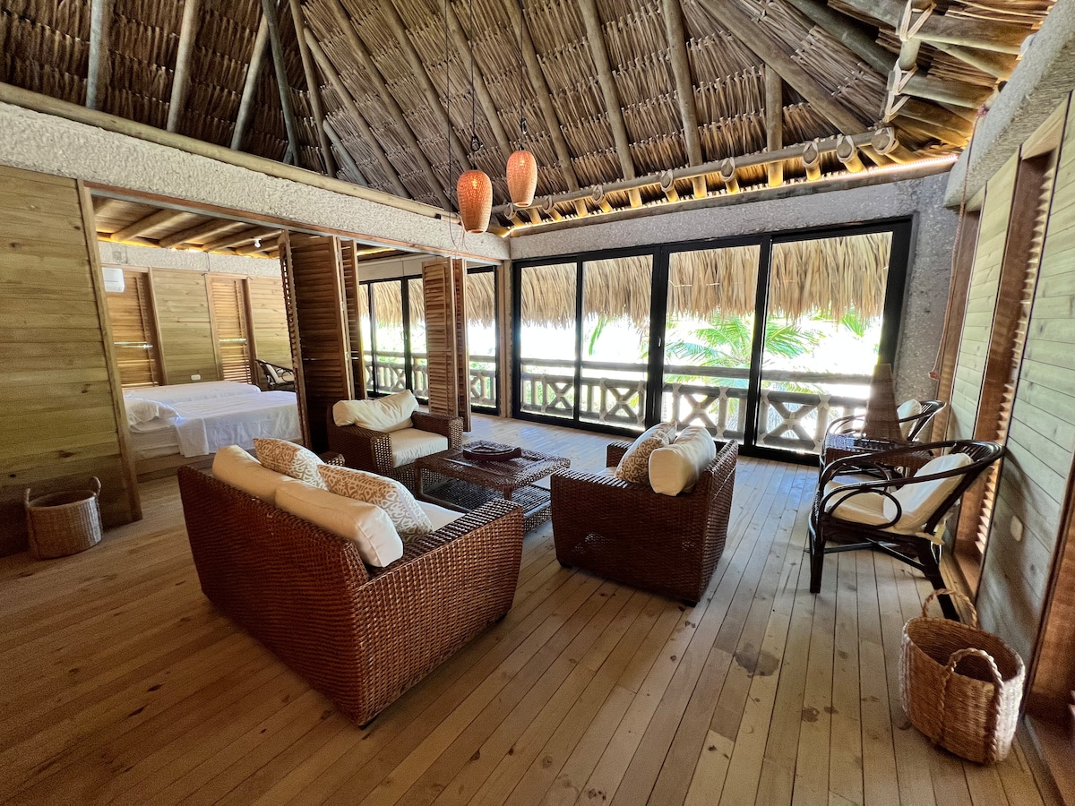 A sunlit seating area features woven chairs and a coffee table surrounded by large windows. A natural wood ceiling with thatched accents complements the warm wooden flooring. The space offers views of the greenery outside, conveying a sense of openness and connection to the outdoors.