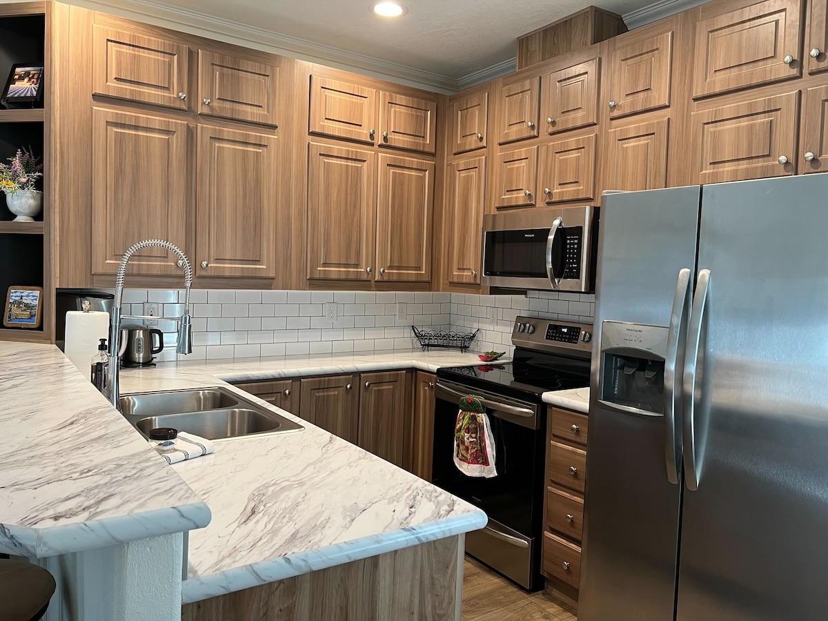 A modern kitchen features wooden cabinets and a marble countertop. Stainless steel appliances include a refrigerator, oven, and microwave. A sleek sink is positioned near the countertop, with a wall-mounted faucet. Subtle white tile backsplashes complement the overall design.