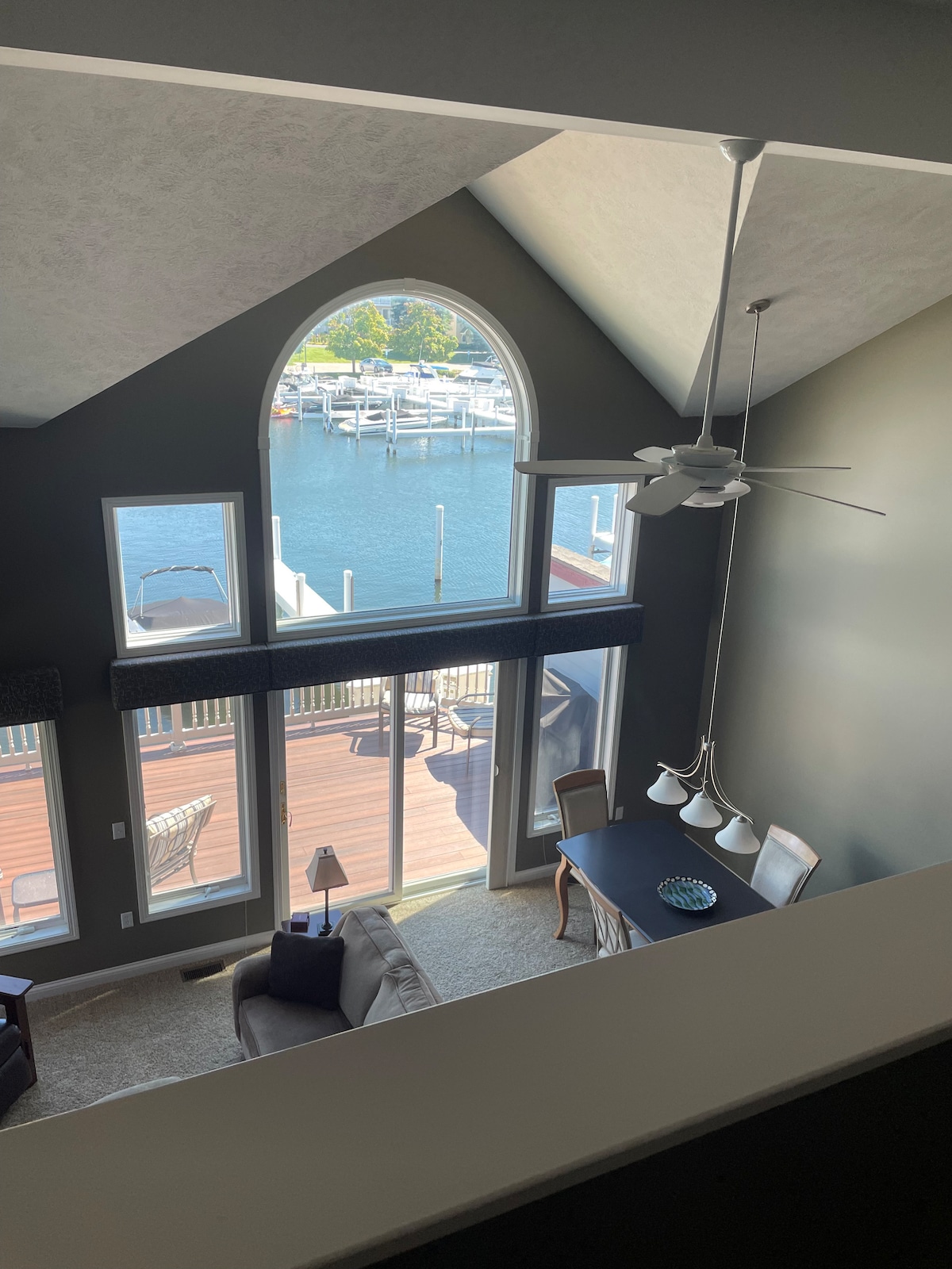 A vaulted ceiling with large arched windows illuminates the open living space. The view showcases the harbor with boats docked, while the dining area is highlighted below, featuring a table with several chairs, a ceiling fan, and soft lighting.