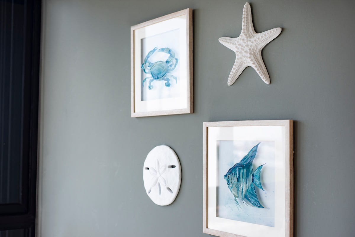 An arrangement of framed marine-themed artwork is displayed on a muted wall. The pieces include an illustration of a crab, a starfish, and a fish, complemented by a circular sand dollar. The artwork adds a coastal charm to the interior.