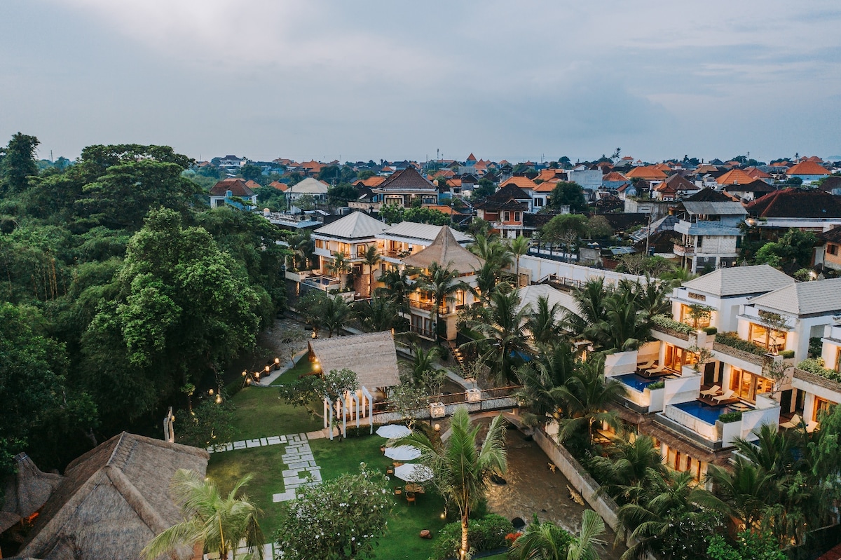 An aerial view of a resort in a lush environment showcases multiple accommodation buildings surrounded by palm trees and manicured gardens. The pathways are lined with soft lighting, creating a serene atmosphere amidst the greenery and neighboring rooftops.