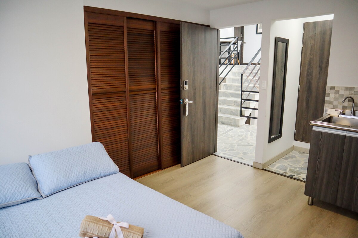 A spacious sleeping area features a neatly made bed with a light blue coverlet. A wooden closet with slatted doors provides storage. The entrance to the bathroom and a small kitchen area are visible, with a staircase leading to another level in the background.