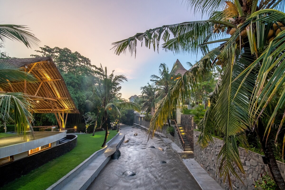 A serene landscape features a gentle river flowing alongside lush greenery and palm trees. A spacious deck area, constructed with natural materials, offers seating while blending harmoniously with the surrounding nature. Soft lighting illuminates the pathway leading to a tranquil setting.