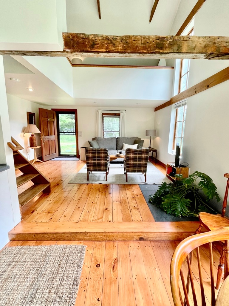 An open living area features warm wooden flooring and high ceilings with exposed beams. Two plaid armchairs sit across from a cozy sofa, accompanied by a small table. Large windows provide views of the surrounding greenery, and a doorway leads to the outdoor space.