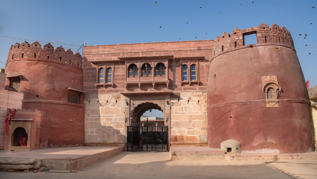 The exterior of a traditional haveli is displayed, characterized by its red sandstone materials and unique architectural features. Towers with crenellated tops flank the entrance, which is adorned with intricately designed arches. The surrounding area is open, providing a sense of space and connection to the environment.