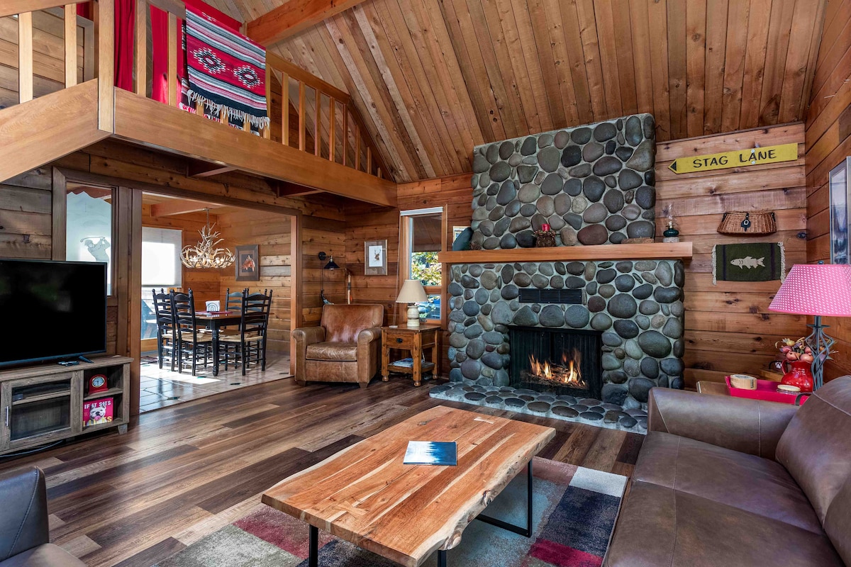 The living room features a stone fireplace set into a wooden wall, surrounded by wood paneling. A flat-screen TV is positioned next to a coffee table, and leather furniture provides seating. A staircase leads to a loft area, visible in the background. Natural light comes from nearby windows.