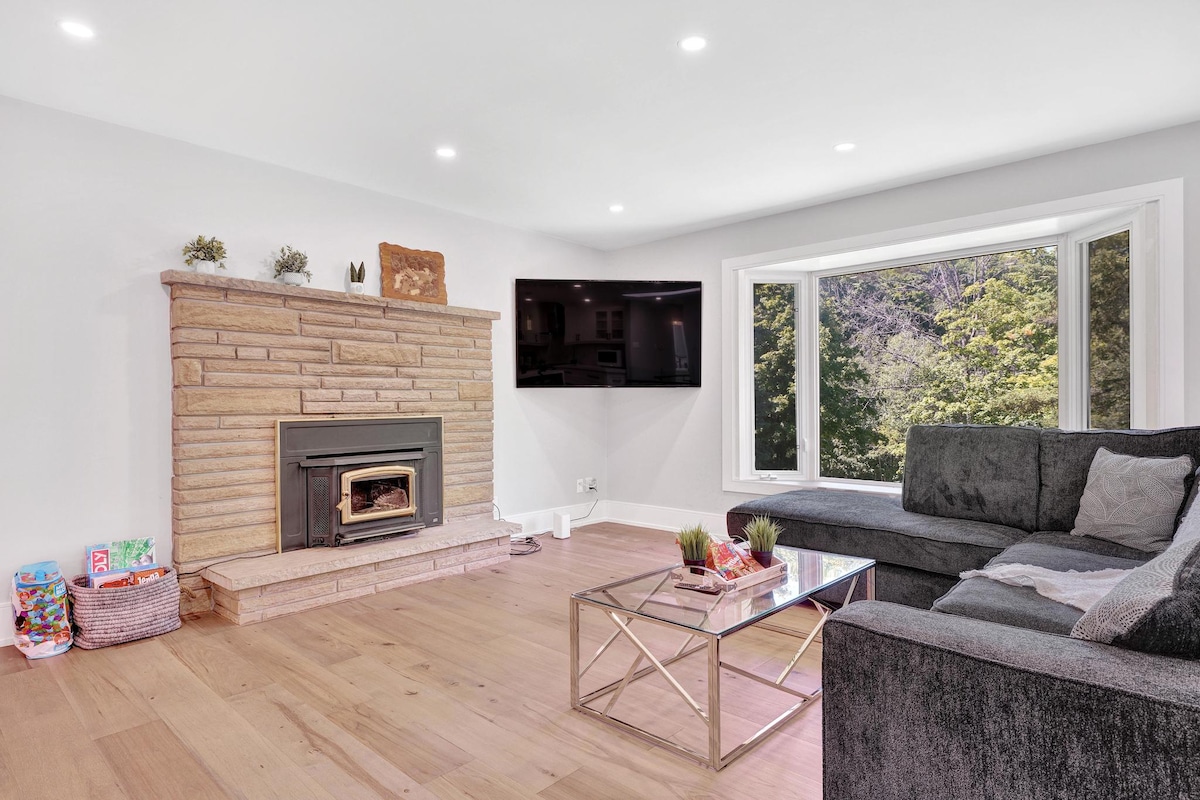 A modern living area features a stone fireplace as a focal point against neutral walls. A large window offers views of the surrounding nature, while a plush sofa and a glass coffee table complete the space. Natural light enhances the warm ambiance.