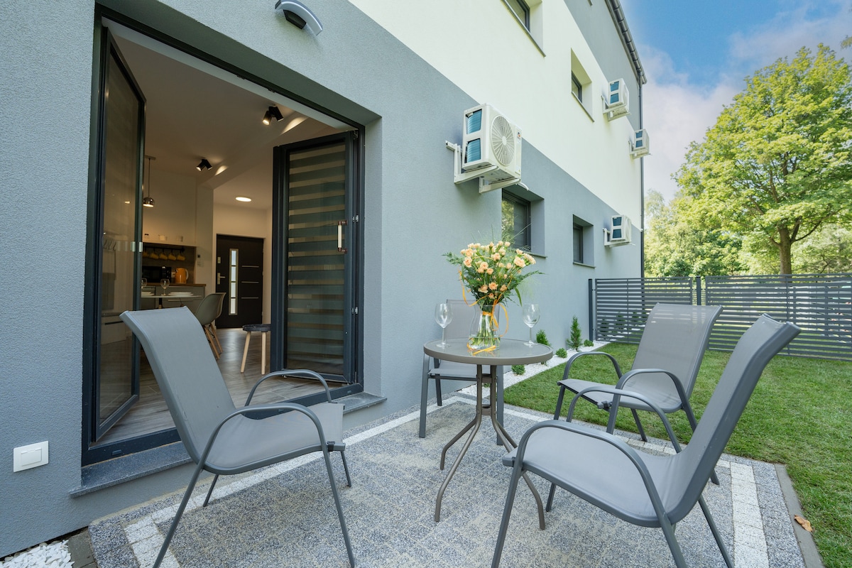 An outdoor seating area is shown with two gray chairs and a small round table. A flower arrangement sits on the table, and a grassy garden is visible in the background, framed by a privacy fence and trees.
