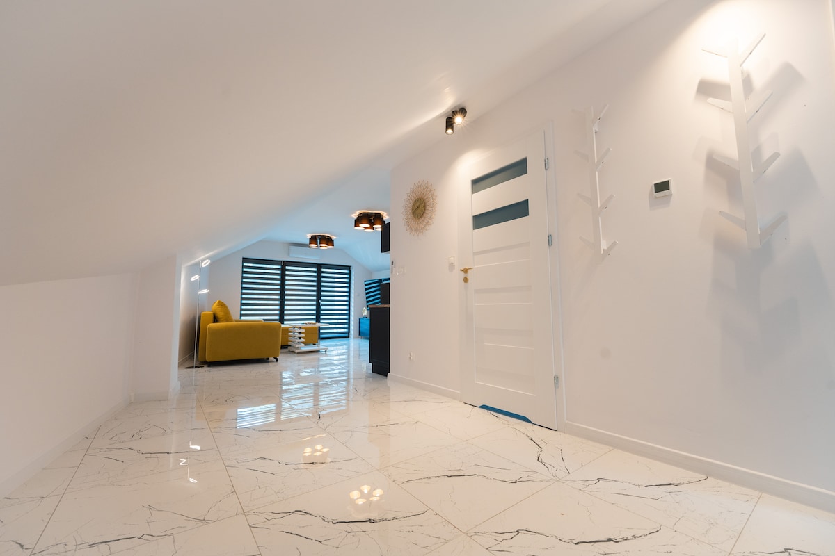 An open living space features shiny marble flooring and white walls. Natural light enters through large windows adorned with black horizontal blinds. A plush yellow sofa adds a pop of color, while modern light fixtures hang softly from the ceiling, complementing the minimalist decor.
