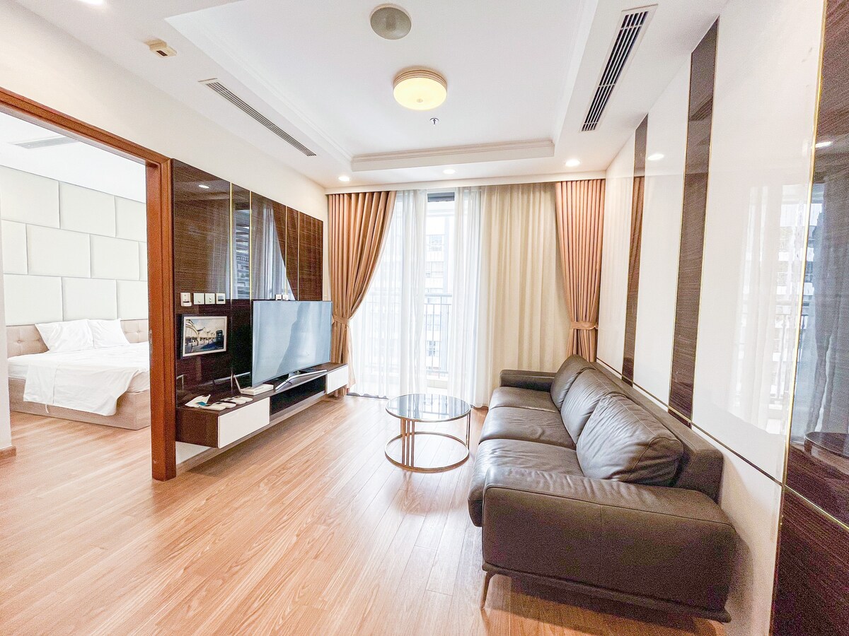 A seating area features a sleek brown leather sofa facing a flat-screen television mounted on an elegant wall unit. Natural light enters through large windows, dressed with light-colored curtains, providing a bright and airy atmosphere. A small round coffee table sits in front, complementing the decor.