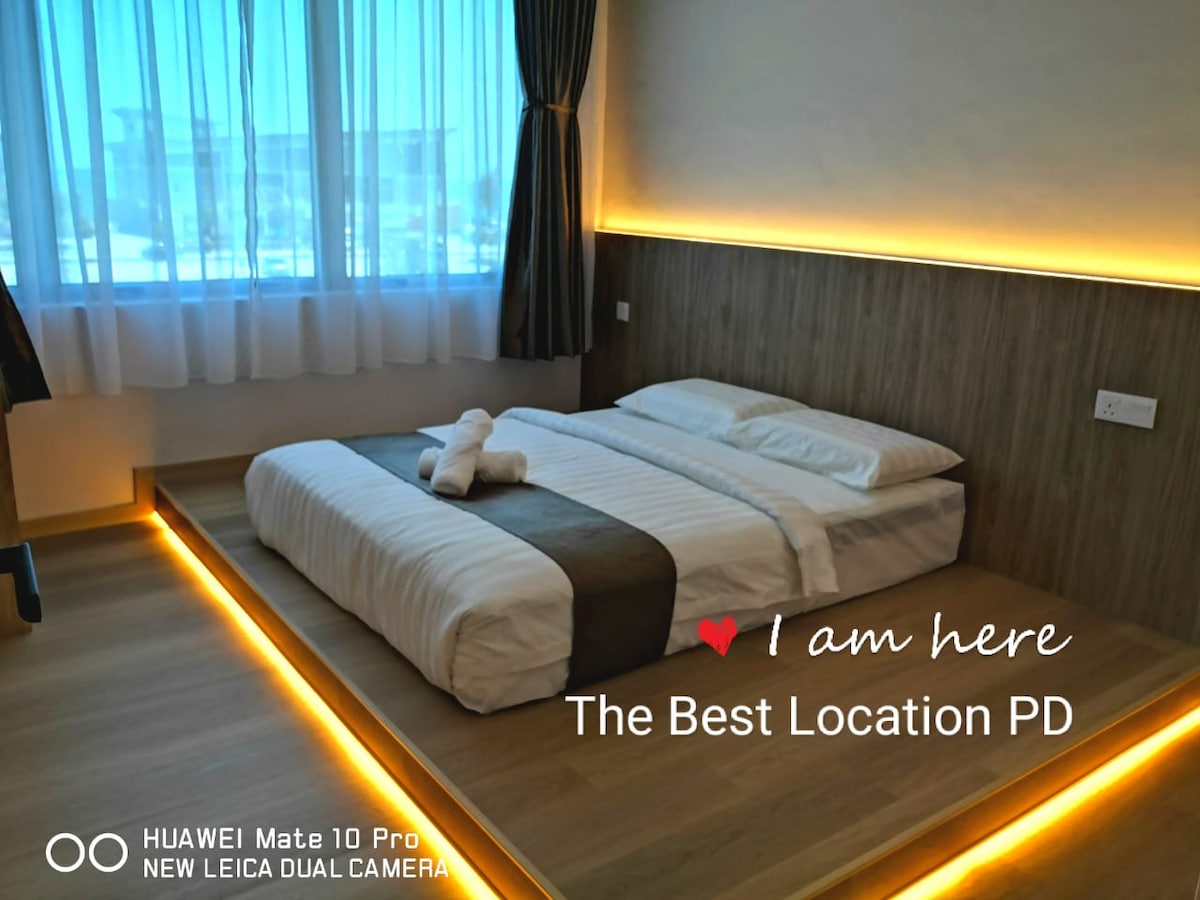 A spacious sleeping area is presented with a low-profile bed featuring light linens and a dark-accented blanket. Soft, ambient lighting encircles the room, complemented by large windows that allow natural light to fill the space, with elegant curtains framing the view.