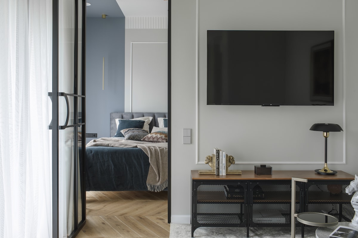 A living area features a sleek, wall-mounted television above a modern media console. A cozy blanket is draped over a nearby chair, while the bedroom is visible through a glass partition, showcasing a spacious bed and soft blue tones.