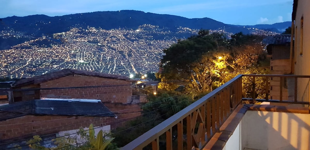 The balcony offers a view of the vibrant city lights sprawling across the hillside, set against a backdrop of distant mountains. A wooden railing frames the space, while nearby trees add a touch of greenery to the picturesque scene.