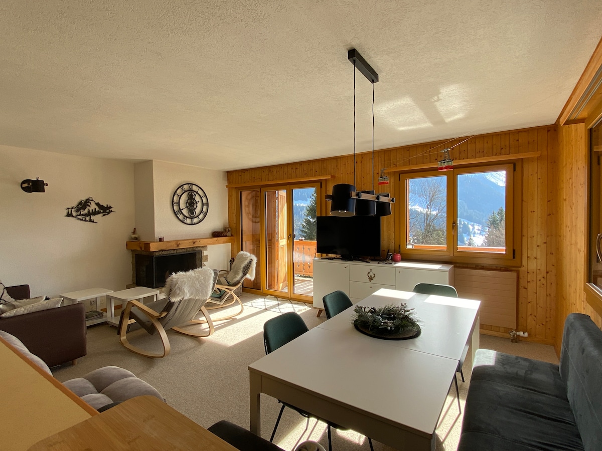 A spacious living area is illuminated by natural light from large windows, offering views of the surrounding mountains. Comfortable seating is arranged around a central table, with a decorative fireplace and a television visible in the background. Wooden accents create a warm ambiance.