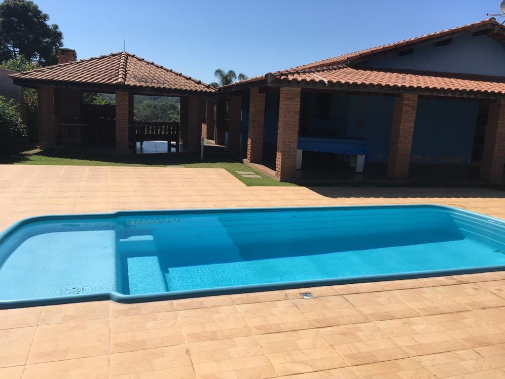 A clear swimming pool is situated in a spacious outdoor area, surrounded by tiled surfaces. Two structures with tiled roofs are visible in the background, providing shaded spaces for relaxation. Lush greenery can be seen in the distance, enhancing the tranquil environment.