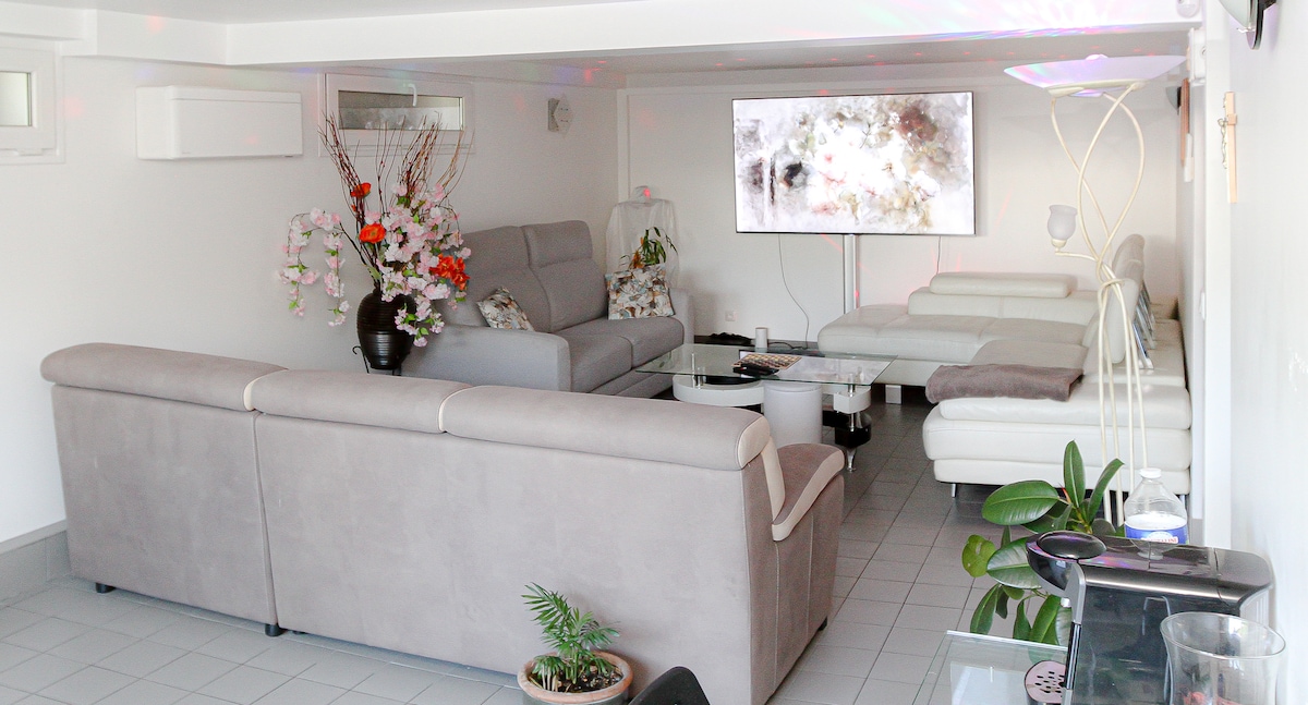 The living area is furnished with a comfortable gray sofa and white sectional seating. A large television mounted on the wall is positioned above a sleek coffee table. Decorative plants and floral arrangements add subtle touches to the bright, open space.