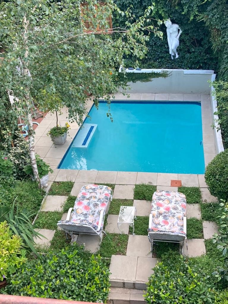 A tranquil outdoor area features a pool surrounded by lush greenery and geometric paving. Two lounge chairs with floral fabric are positioned beside the pool, offering a serene spot for relaxation. A statue can be seen among the greenery, enhancing the peaceful atmosphere.