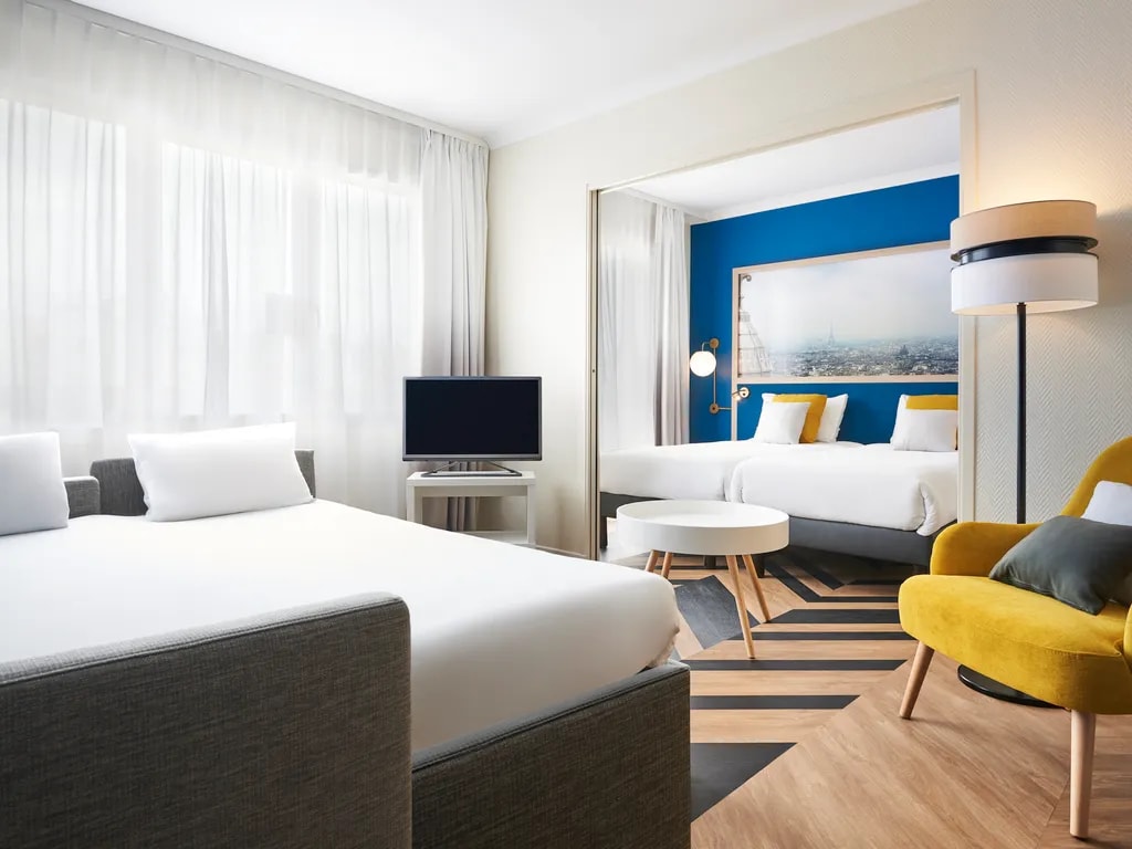A well-lit guest room features a spacious bed and a bright blue accent wall. A mirror reflects a second sleeping area with a large bed. A comfortable yellow chair and a small round table complement the modern decor. Natural light filters through sheer curtains.