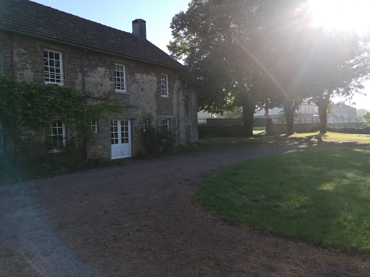 The exterior of the stone house is framed by lush greenery, with a gravel pathway leading to the entrance. Natural light filters through tall trees, casting soft shadows on the ground. The spacious yard features a mix of grassy areas and gravel.