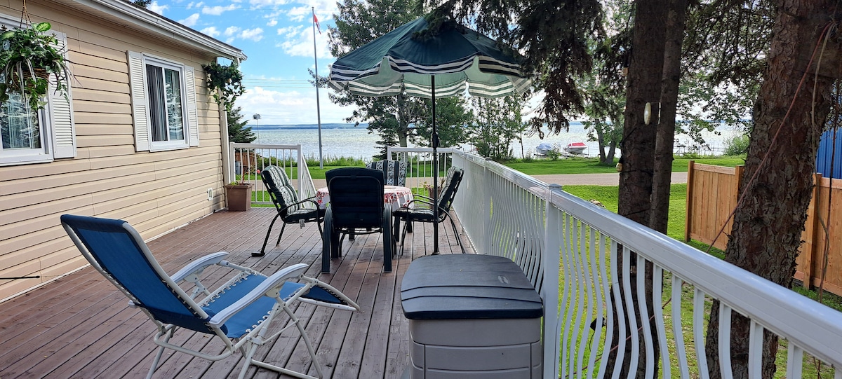 A spacious deck area features several chairs arranged around a table, complemented by a patio umbrella. Lush greenery surrounds the decking, with a clear view of the lake visible in the background. A storage box is positioned nearby, providing additional convenience.