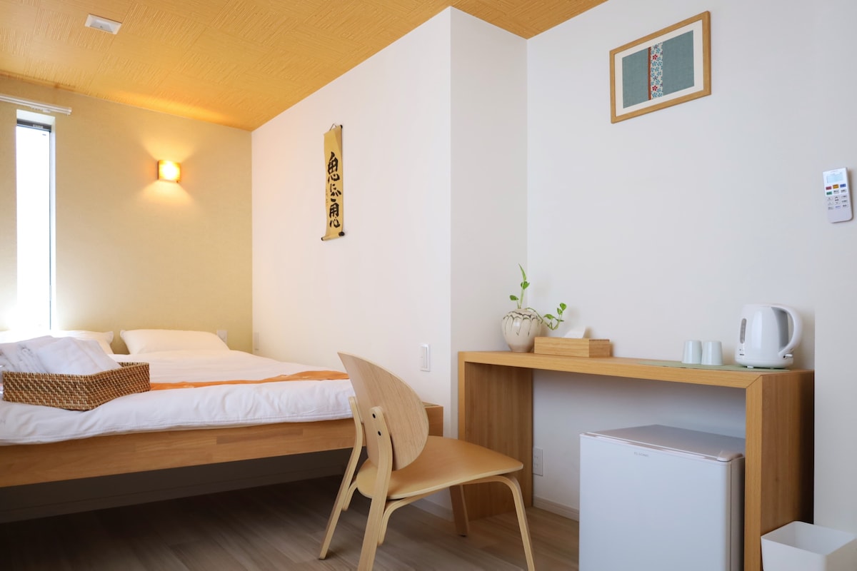 The room features a double bed with crisp linens, accompanied by a wooden desk and a simple chair. A refrigerator and kettle are visible beside the desk. Natural light enters through a window dressed with light curtains, and minimal decor adds a serene touch.