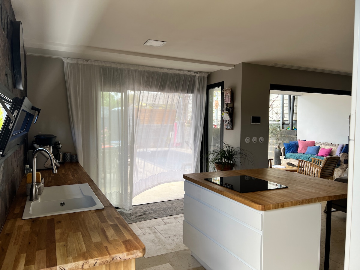 A modern kitchen is visible, featuring a wooden countertop and a sleek island with an integrated cooktop. Sheer curtains allow natural light to filter in, connecting the space to an outdoor area that can be glimpsed through glass doors.