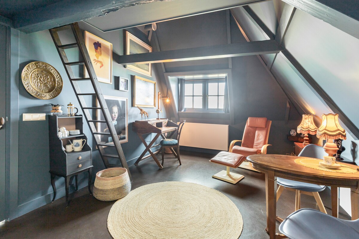 A cozy studio space is highlighted by warm, muted colors and a mix of vintage decor. A small wooden table and two chairs are accompanied by a lounge chair and a desk. A ladder leads to a lofted sleeping area, with light filtering through the window.