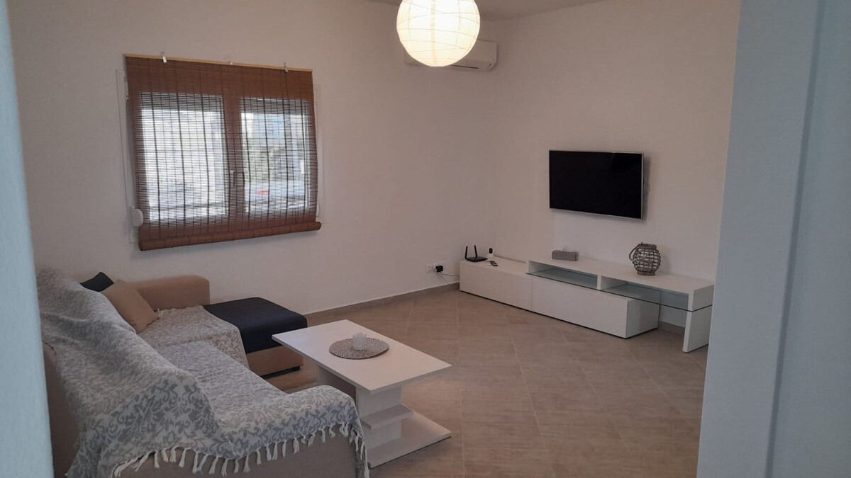 A spacious living area is highlighted by a light-colored sofa and a contrasting dark accent piece. A modern television is mounted on the wall, with a minimalist white coffee table in front. Natural light enters through the window covered with bamboo shades.