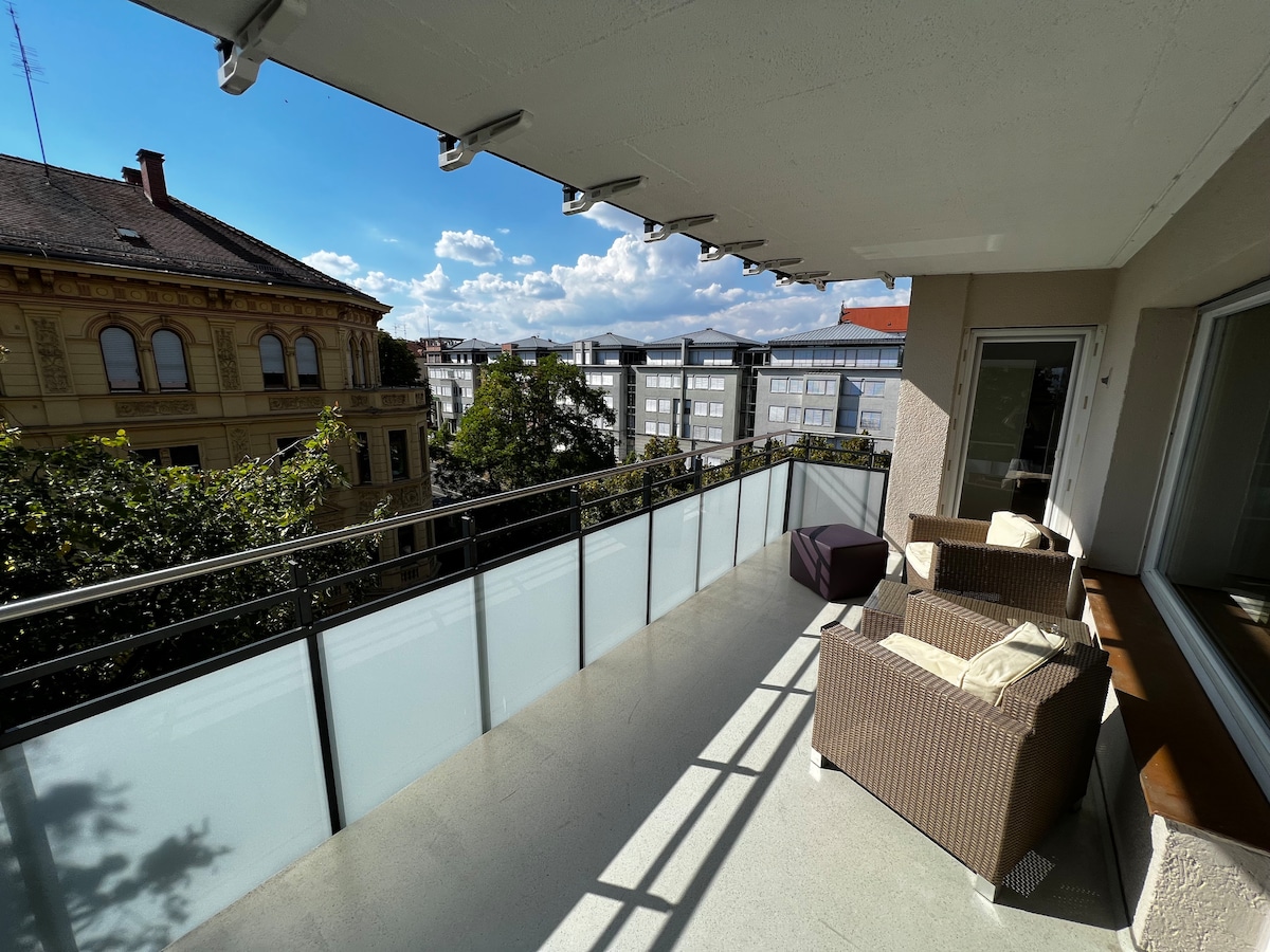 A spacious balcony features a seating area with wicker furniture, overlooking nearby buildings and green trees. The glass railing allows for unobstructed views of the surroundings and natural light. The area is partially shaded, creating a comfortable space for relaxation.
