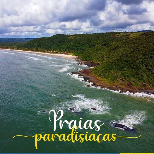 A scenic coastal view reveals a calm ocean meeting sandy beaches bordered by lush green hills. Gentle waves create a soft foam along the shore, while clouds drift in a partly blue sky, highlighting the tranquility of the beach environment.