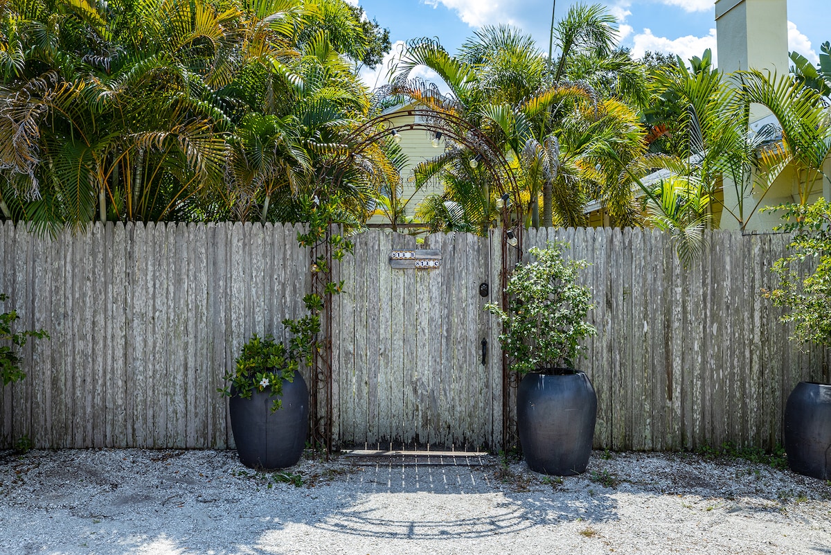 The entrance gate is framed by lush tropical foliage, creating a sense of privacy. A weathered wooden fence offers a rustic charm, while two large planters with greenery stand on either side of the path leading into the property.