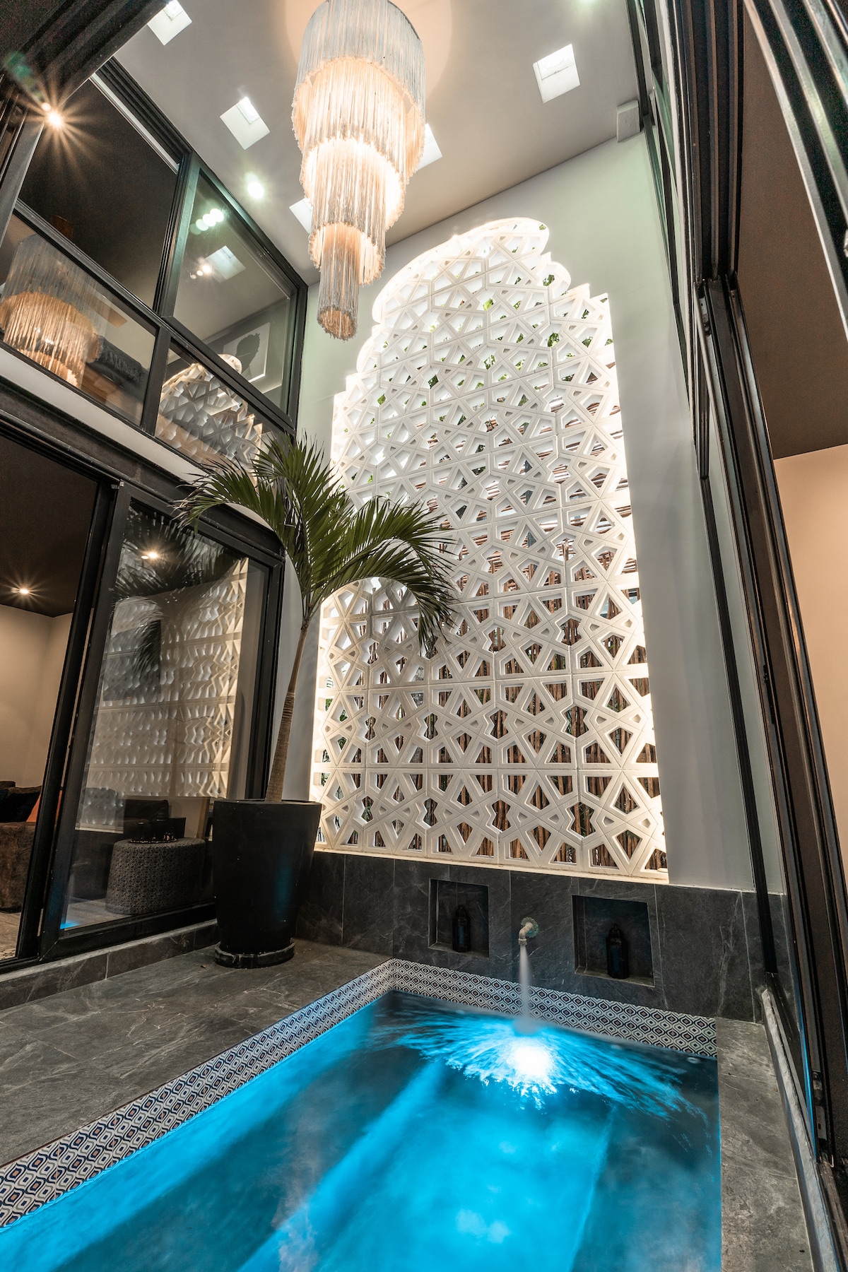 A private plunge pool is illuminated with soft blue lighting, surrounded by sleek gray tiles. A tall decorative wall features intricate geometric patterns, complemented by a stylish chandelier overhead. A palm plant adds a touch of greenery to the serene setting.