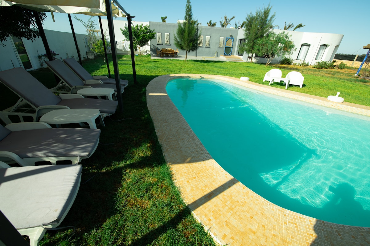 A tranquil swimming pool is surrounded by a grassy area, complemented by several loungers. The pool's clear water reflects the blue sky, while a spacious outdoor area with trees and a seating area can be seen in the background.