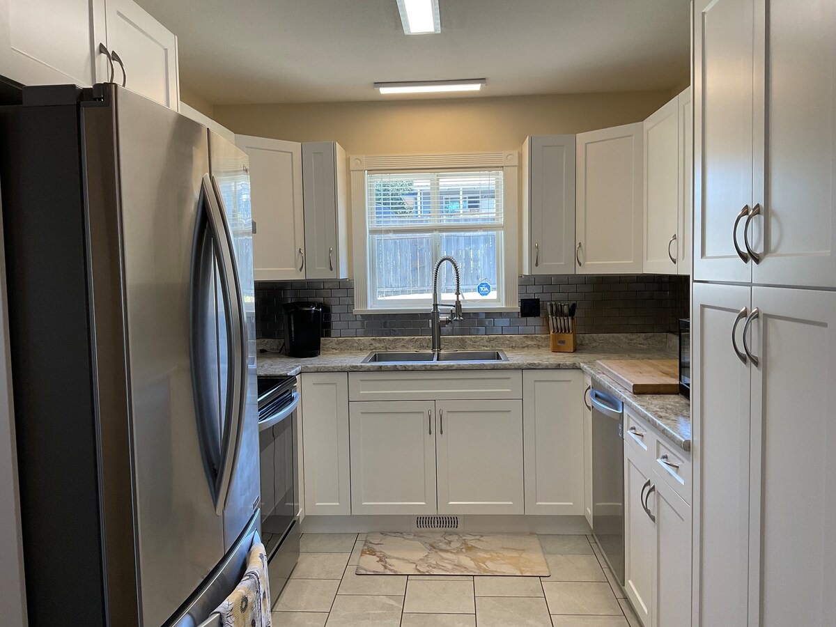 A modern kitchen features white cabinetry and granite countertops. Stainless steel appliances, including a refrigerator and oven, are positioned conveniently. A large window allows natural light to illuminate the space, while a sink is centrally located beneath the window, enhancing functionality.