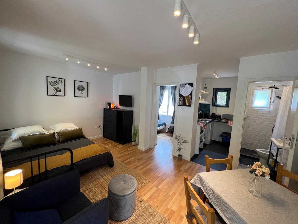 An open living space features a cozy area with a stylish bed and a sofa, alongside a dining table with four chairs. A fully equipped kitchen is visible, with natural light filtering in through a window. The decor includes minimalistic wall art and a welcoming layout.