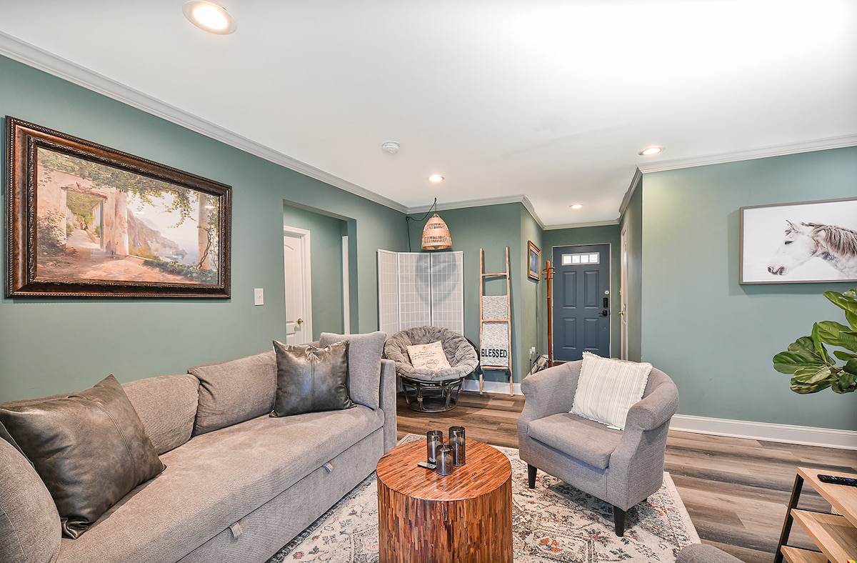 A cozy living area features a soft gray sofa and an accent chair, with a wooden coffee table positioned at the center. The walls are painted in a calming green, complemented by decorative art and plants, creating an inviting atmosphere. A front door and hallway can also be seen.