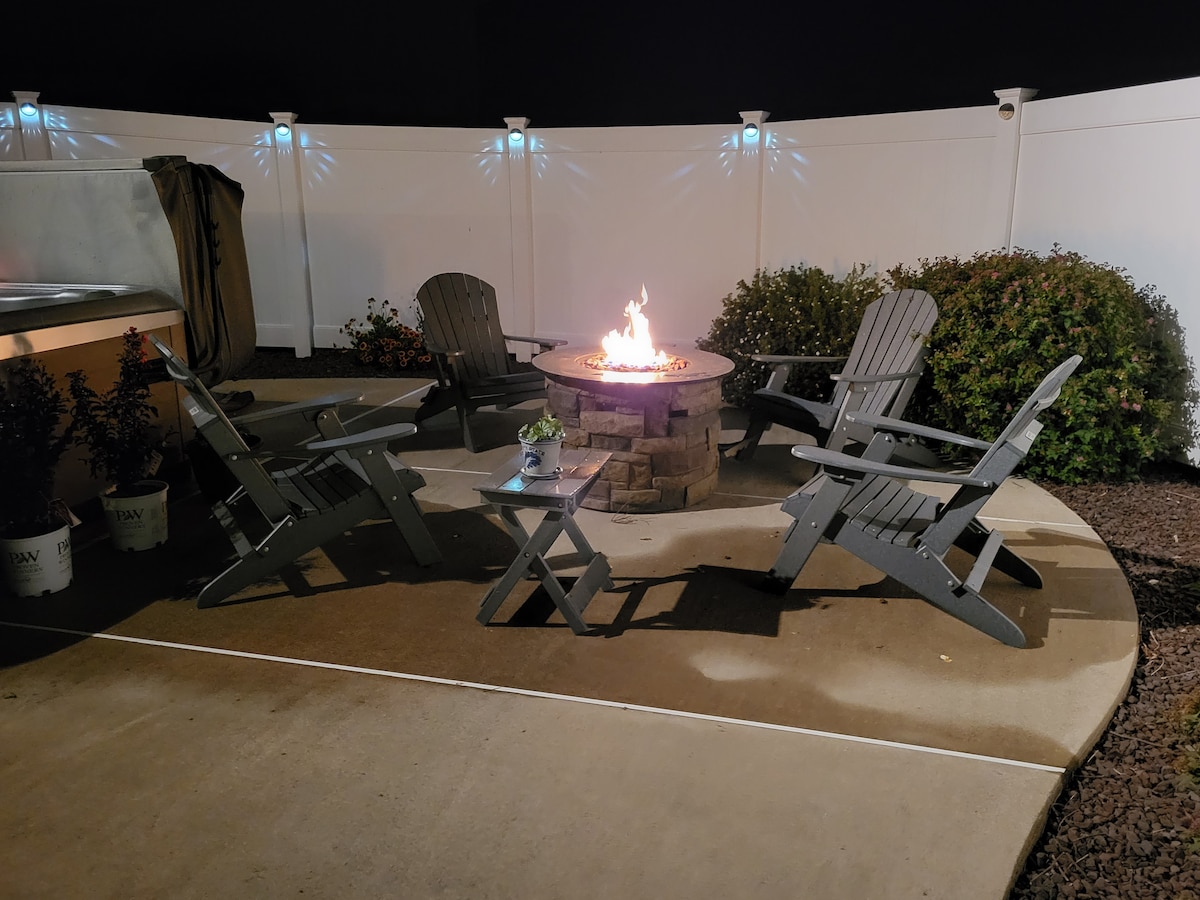A cozy outdoor seating area features four gray Adirondack chairs arranged around a stone fire pit. A small side table rests nearby, illuminated by the warm glow of the flames. Green shrubs line the background, enhancing the inviting space.