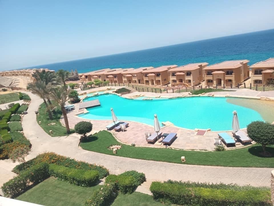 A scenic view showcases a large, vibrant pool framed by lush greenery and elegant lounge chairs. The turquoise water reflects the clear blue sky and is complemented by nearby sandy shores and residential buildings, creating a tranquil coastal atmosphere.