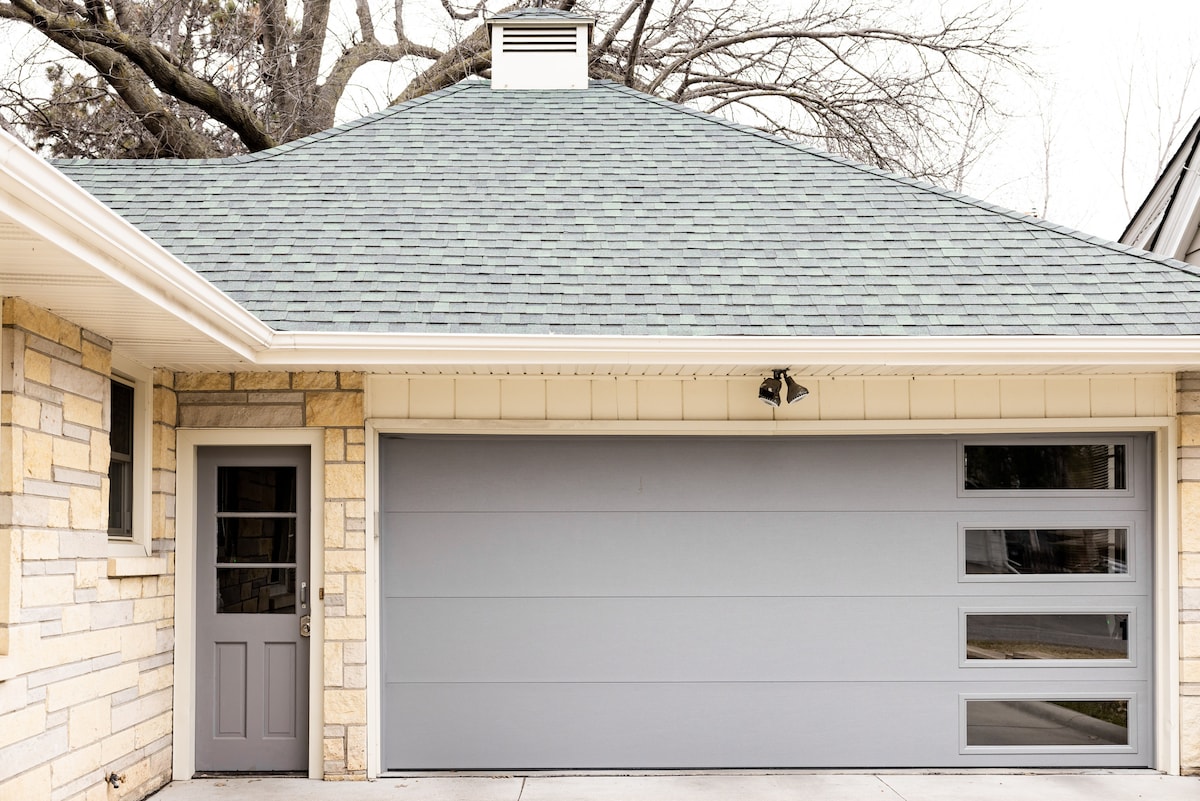 The exterior of the home features a stone façade complemented by a sleek gray garage door showcasing three clear window panels. A side door with glass panels is visible, enhancing the modern aesthetic. The roof is structured in a gentle slope, adorned with shingles.