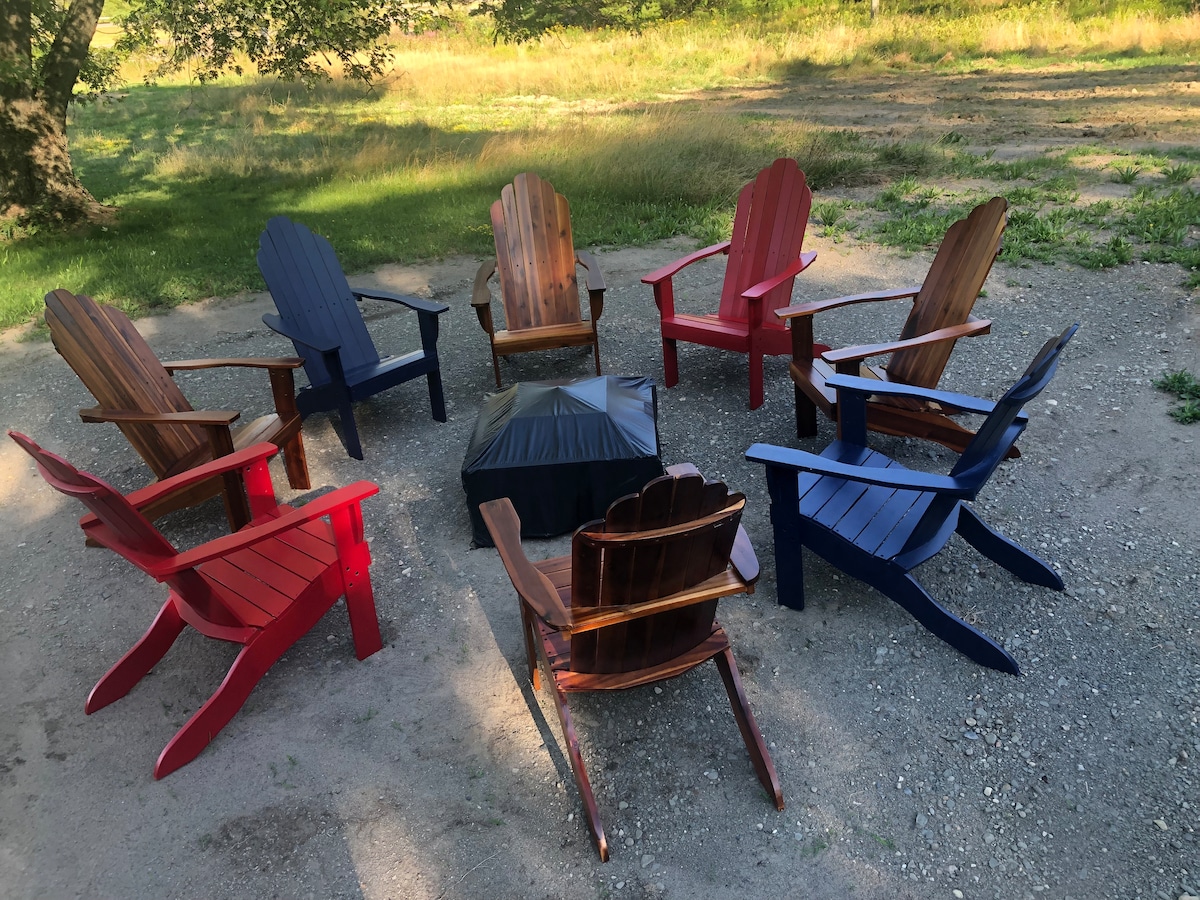A circular arrangement of ten Adirondack chairs in varying colors, including red, blue, and natural wood tones, surrounds a fire pit. The chairs are set on a gravel surface in a grassy area, creating an inviting outdoor gathering space.