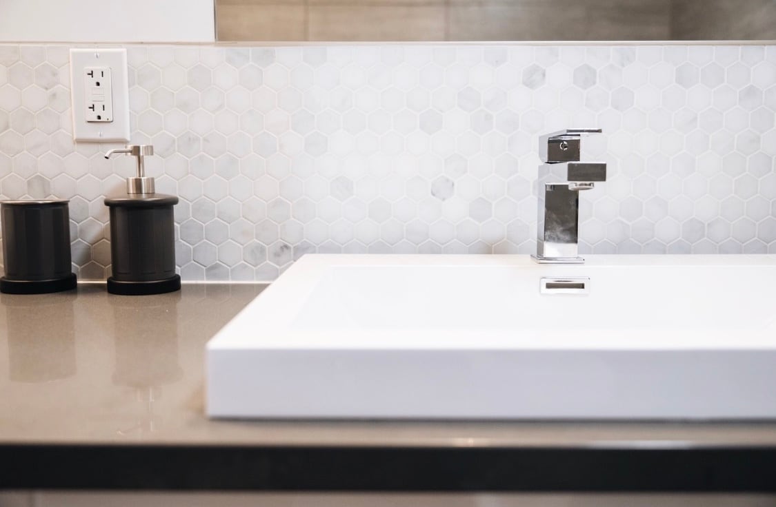 A modern bathroom sink is displayed with a sleek faucet positioned above a white rectangular basin. The countertop features a polished dark surface, complemented by a decorative hexagonal tile backsplash. Two small black containers are placed beside the sink for storage.